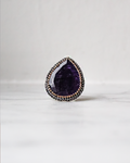 A teardrop-shaped purple gemstone ring with a silver-toned halo accented by small rose gold stones, resting on a light marble surface against a white background.