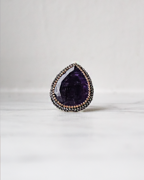 A teardrop-shaped purple gemstone ring with a silver-toned halo accented by small rose gold stones, resting on a light marble surface against a white background.