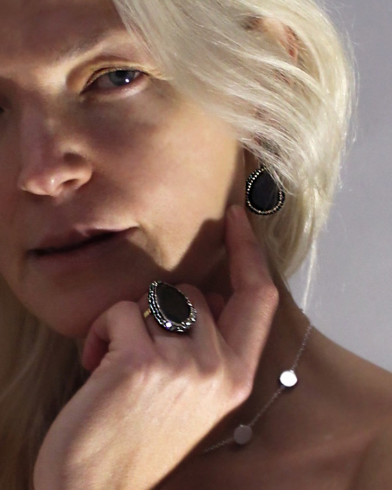 A close-up lifestyle shot of a blonde woman wearing a matching jewelry set, featuring a large oval amethyst stone ring on her finger and a matching teardrop-shaped earring, with a delicate silver chain necklace.