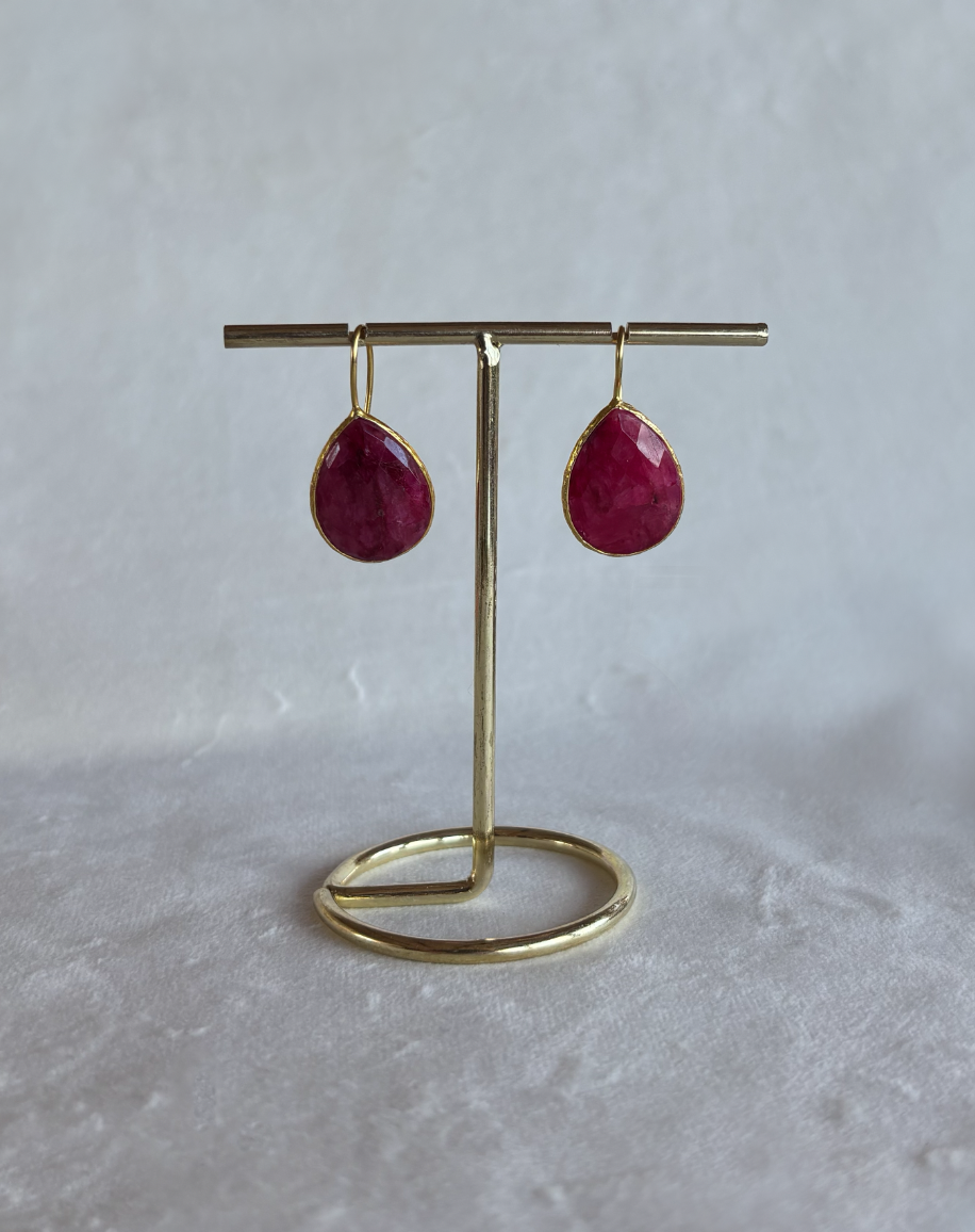 A pair of gold teardrop dangle earrings with deep red gemstones, displayed on a gold stand against a textured light gray background.