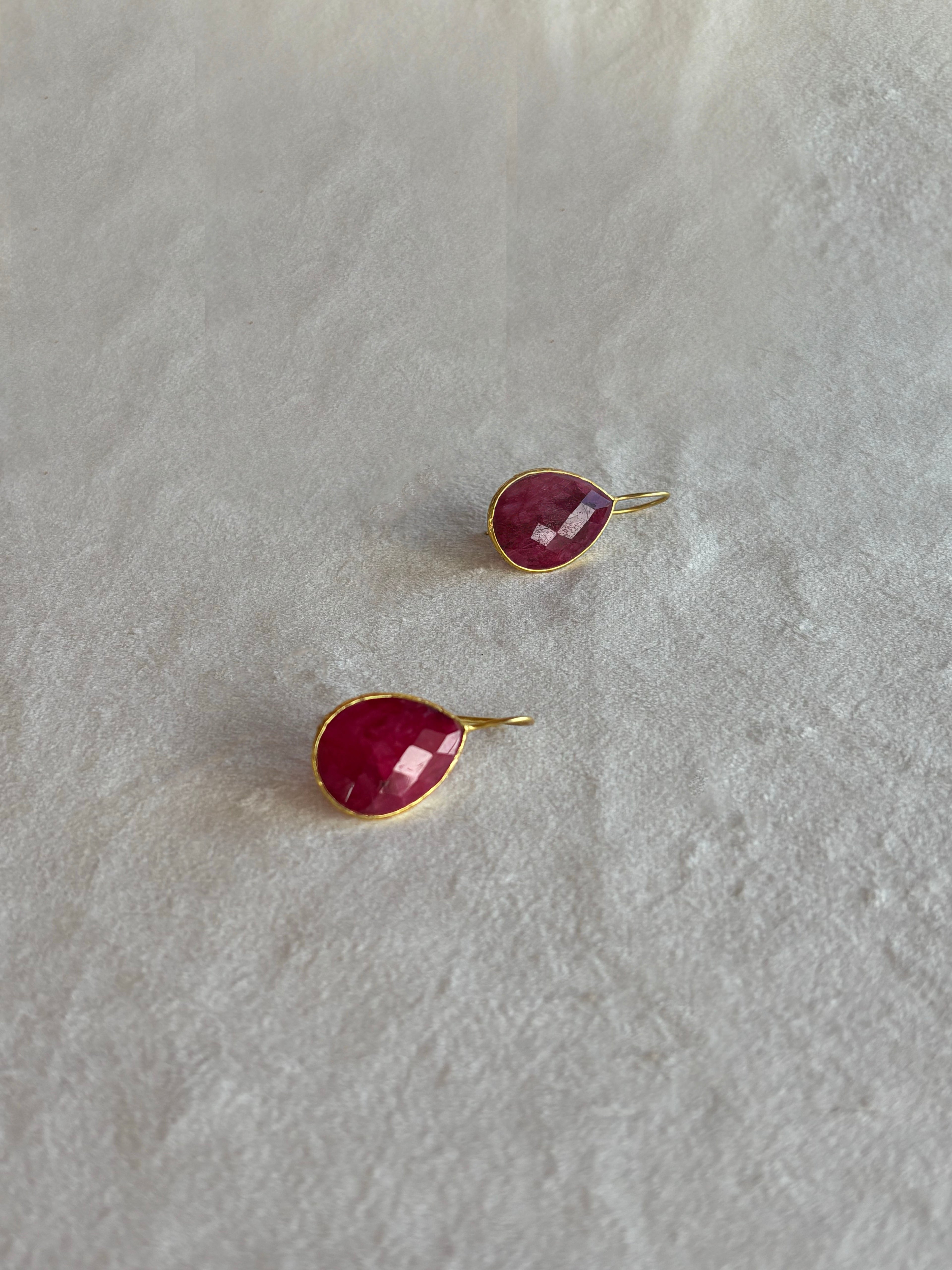 A pair of gold hoop earrings with oval-cut deep pinkish-red cabochon gemstones, displayed on a textured white surface.