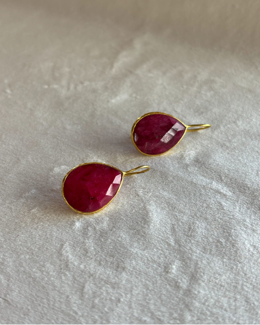 A pair of gold teardrop-shaped ruby earrings with faceted deep pinkish-red gemstones, resting on a textured off-white fabric background.