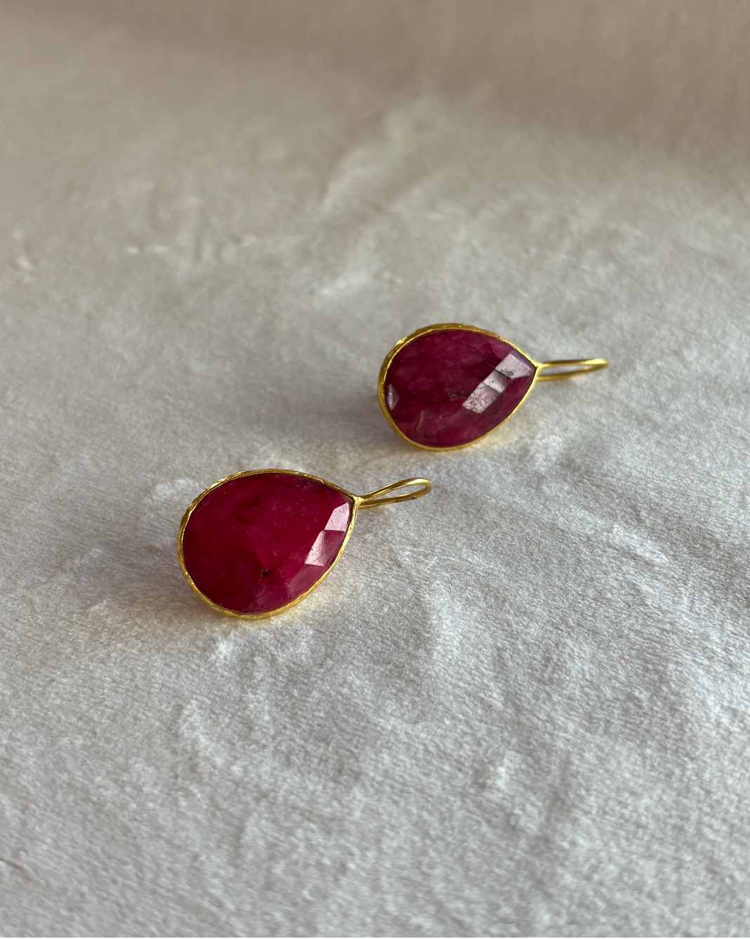 A pair of gold teardrop-shaped ruby earrings with faceted deep pinkish-red gemstones, resting on a textured off-white fabric background.