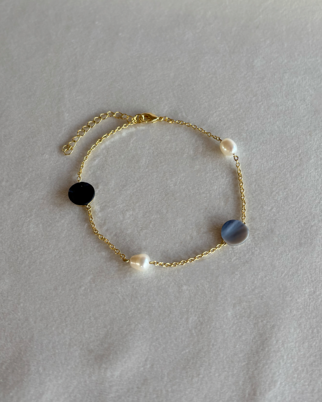 A beautiful gold chain bracelet featuring  blue mussel  mother of pearl in light blue and marine blue hues and fresh water pearls. round.textured white fabric background.